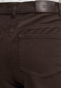 Pantalon en denim brun foncé vu de dos montrant une poche unique et un patch en cuir de marque portant l'étiquette "ONLY Since 1995" sur la ceinture.