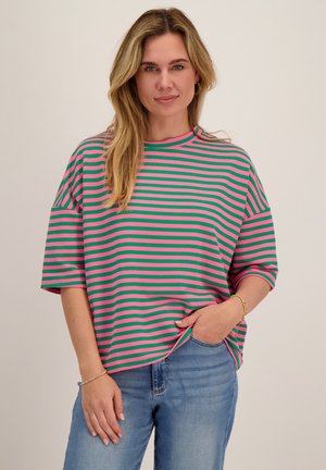 Vrouw die een oversized T-shirt met groene en roze horizontale strepen draagt en een blauwe spijkerbroek, poseert met één hand in de zak tegen een effen achtergrond.