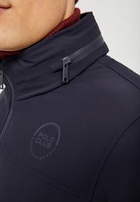 Navy jacket with a high collar, textured fabric, zippered pockets, and a printed logo detailing "POLO CLUB ESTABLISHED 2012" on the chest.