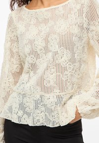 Kvinne iført en kremfarget blondetopp med lange ermer, blomsterdesign og volangkant, kombinert med svarte bukser.