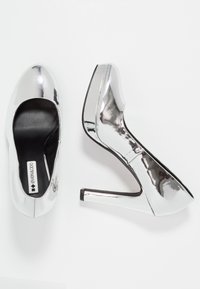 Srebrne metaliczne buty na wysokim obcasie z platformą, jeden widoczny z góry, a drugi z boku, na białym tle.