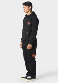 Sudadera negra con capucha y bolsillo frontal, con un diseño gráfico en rojo; combinada con pantalones cargo negros que presentan acentos rojos. Calzado informal.