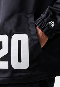 Black jacket with white "20" and contrasting stitching, featuring a side pocket and elastic cuffs for a fitted design.
