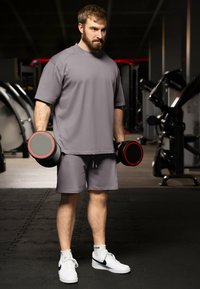 Maglietta atletica grigia e pantaloni corti, che tiene in mano manubri neri e grigi con accenti rossi. Sneakers bianche con swoosh nero su un pavimento della palestra.