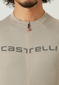 Ensemble de cyclisme beige avec une fermeture éclair mi-longue, arborant un logo "castelli" gris sur la poitrine et une texture de tissu lisse et extensible.
