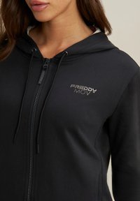 Sudadera negra con cremallera confeccionada en una tela suave. Cuenta con una capucha, cordones y un logo plateado en el pecho. Detalle de superficie texturizada visible.