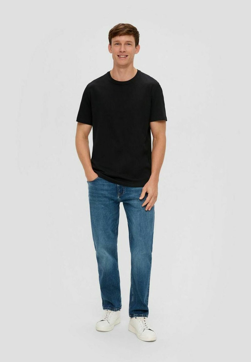 Schwarzes Baumwoll-T-Shirt mit kurzen Ärmeln, kombiniert mit hellblauen Jeans und weißen Sneakers. Das Model steht vor einem schlichten Hintergrund.