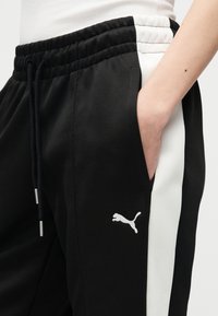 Κοντινό πλάνο ενός ατόμου που φοράει μαύρο παντελόνι φόρμας Puma με λευκές πλευρικές ρίγες και το χέρι μέσα στην δεξιά τσέπη.