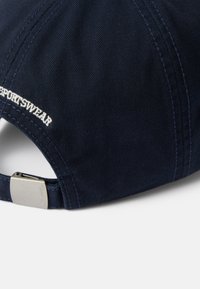 Tummansininen baseball-lippalakki, joka on valmistettu puuvillasta. Siinä on kontrastiväriset saumat, valkoisella brodeerattu "SPORTSWEAR" teksti ja hopeanvärinen metalli solki takana.