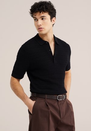 Junger Mann mit lockigem Haar, der ein strukturiertes schwarzes Kurzarm-Poloshirt und braune Hose mit schwarzem Gürtel trägt, Hände in den Taschen, schaut nach links.