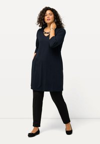 Marineblaue Tunika mit V-Ausschnitt und kreuzförmigen Details, kombiniert mit schwarzen, eng anliegenden Hosen und schwarzen Ballerinas. Aus weichem, glattem Stoff gefertigt und mit seitlichen Taschen.