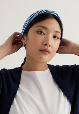 Ung kvinde justerer blåt og hvidt stribet pandebånd, iført guldøreringe, hvid skjorte og mørkeblå cardigan mod en ensfarvet baggrund.