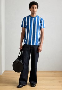 Camisola de manga curta às riscas azul e branca, jeans largos e escuros, segurando uma mala redonda preta, de pé sobre um piso de madeira contra um fundo neutro.