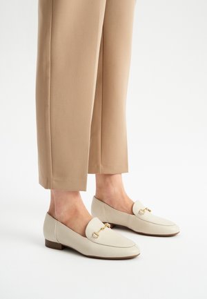 Beige nahkakengät, joissa on suorakulmainen kultainen yksityiskohta, matala lohkokorko ja sileä pinta, yhdistettynä beigeihin kapeneviin housuihin.