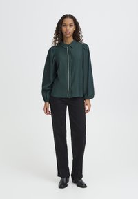 Blusa verde texturizada con cuello y cierre de botones delanteros, combinada con pantalones negros de pierna recta y botas negras de tacón.