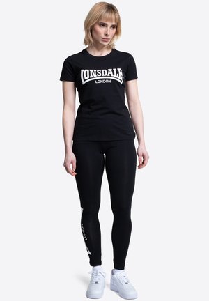 Žena stojaca, oblečená v čiernom tričku "Lonsdale London", čiernych legínach s bielym textom na jednom stehne a bielych teniskách.