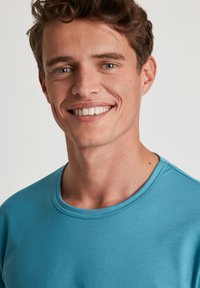 Turquoise sweatshirt met een ronde hals, zachte textuur en subtiele naaddetails. Glad weefsel valt comfortabel om de hals en schouders.