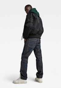 Zwarte bomberjack, groene hoodie eronder, donkere spijkerbroek met zichtbare stiksels, en witte sneakers. Achteraanzicht, neutrale achtergrond.