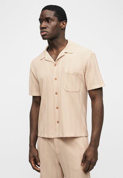 Jeune homme noir portant une chemise beige à manches courtes avec une poche et un pantalon assorti, debout devant un fond blanc uni.