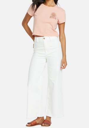 Mujer con camiseta corta de color rosa claro con gráfico de sol, pantalones blancos de cintura alta y pierna ancha, y sandalias marrones con los dedos al aire.