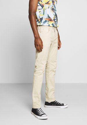 Man draagt beige rechte pijpen broek, tropisch patroon korte mouwen overhemd en zwarte hoge sneakers, staand tegen een effen achtergrond.