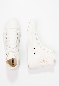 Zapatillas altas blancas de material de lona, diseño con cordones, puntera de goma y parche redondo con logo en el lateral. Textura suave y líneas limpias.