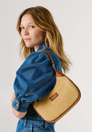 Mujer joven con cabello rubio hasta los hombros, vistiendo una chaqueta vaquera y llevando un bolso tejido color marrón claro con detalles de cuero marrón sobre el hombro.