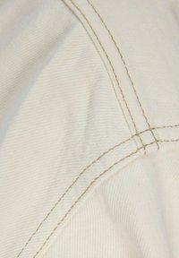 Κοντινή λήψη κρεμ υφάσματος με ορατές διπλές ραφές μπεζ χρώματος που διασχίζουν διαγώνια και συναντώνται σε μια γωνία.