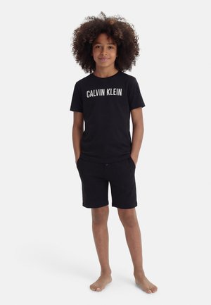 Otrok z zvitimi lasmi, oblečen v črno majico Calvin Klein in ujemajoče se črne kratke hlače, stoji bos z rokami v žepih.