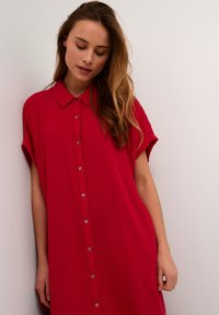 Abito camicia rosso, con bottoni e colletto, maniche corte e tessuto strutturato. Presenta una vestibilità ampia e un sottile dettaglio dei bottoni.