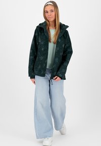Donkergroene jas met een sterpatroon, lichtgewicht materiaal, verstelbare capuchon, gecombineerd met lichtblauwe wijde jeans en witte sneakers.