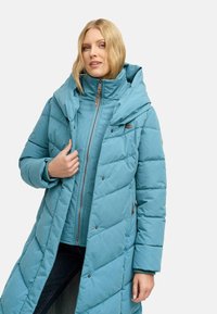 Długi tealowy płaszcz puchowy z kapturem, z ukośnym przeszyciem, zatrzaskami i zamkiem błyskawicznym z przodu, nałożony na pasującą kurtkę.