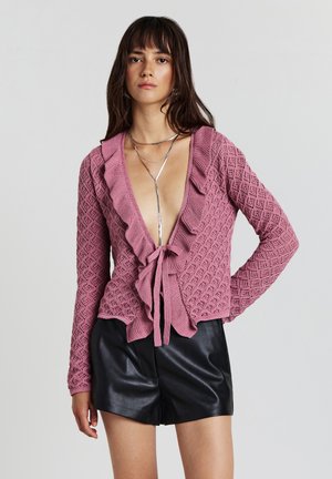 Cardigan en tricot rose doux avec encolure à volants, fermeture à nouer et motif en losanges. Assorti à des shorts en cuir noirs.