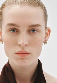 Modelo con cabello lacio y claro, usando pendientes de aro dorados. El enfoque está en su rostro, con maquillaje mínimo y una bufanda de color marrón oscuro.