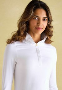 Weißes Langarmoberteil mit einem geknöpften Kragen, das eine satinartige Textur und eine Spitze entlang des Saums aufweist. Angenehme Passform aus weichem Stoff.