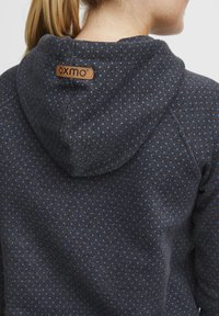 Tamno plava dukserica s kapuljačom, ukrašena malim svijetloplavim točkicama, s kožnom etiketom "OXMO" na zadnjoj strani. Mekan, teksturiran materijal.