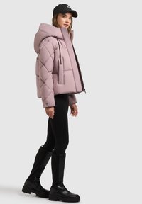 Svetloružová nadýchaná bunda s vysokým golierom, prešívaným vzorom v tvare diamantu, predným zipsom a bočnou kapsou, skombinovaná s čiernymi legínami a čižmami po kolená.