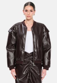 Giacca bomber in ecopelle marrone con polsini e colletto a coste, caratterizzata da una chiusura frontale con zip e silhouette oversize. Texture liscia e dettagli cuciti.