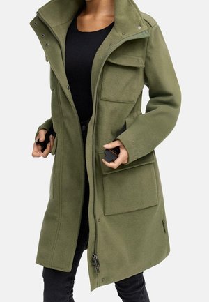 Woman wearing an open olive green fleece coat with large front pockets over a black top and black pants, holding black objects in both hands.