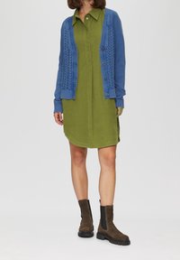 Cárdigan de punto azul con patrón de trenzas, llevado sobre un vestido verde con cuello. Botines de ante marrón con una suela negra gruesa.