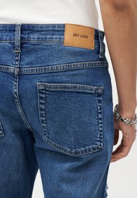 Άτομο που φοράει μπλε τζιν τζιν με δερμάτινη ετικέτα στην μέση με την επιγραφή "ONLY & SONS" και ασημένιο βραχιόλι με αλυσίδα στον καρπό.