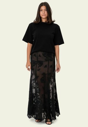 T-shirt noir à manches courtes associée à une jupe longue noire transparente ornée de motifs en dentelle florale et d'un ourlet festonné ; le tout porté avec des sandales noires.