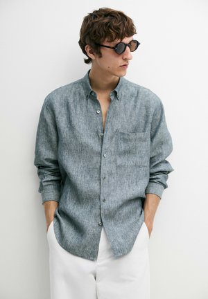 Jeune homme portant des lunettes de soleil noires rondes, une chemise grise boutonnée avec poche, et un pantalon blanc, debout les mains dans les poches.