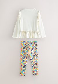 Witte top met lange mouwen, gerimpelde schouders en een peplumzoom, gecombineerd met meerkleurige legging met een regenboog- en sterrenpatroon op een donkere achtergrond.