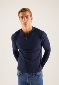 Cămașă henley cu mâneci lungi, de culoare bleumarin, cu guler rotund, nasturi cu trei butoniere și manșete striate, asortată cu blugi albaștri. Textură netedă.