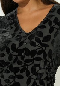 Czarna sukienka z teksturowanym wzorem liści, dekoltem w kształcie v oraz guzikami z przodu. Materiał wydaje się miękki i lekko refleksyjny.