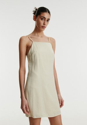 Cremfarbendes Minikleid mit dünnen Trägern, geradem Ausschnitt und lockerer Passform. Hergestellt aus leichtem Stoff mit glatter Textur.