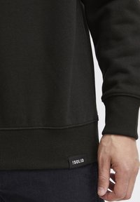 Sweatshirt noir en tissu doux avec des poignets et un ourlet côtelés. Présente une petite étiquette avec « !SOLID » en bas à gauche.