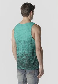 Hombre de espaldas con camiseta sin mangas texturizada en turquesa y negro y pantalones cortos vaqueros azules descoloridos, frente a un fondo claro y liso.