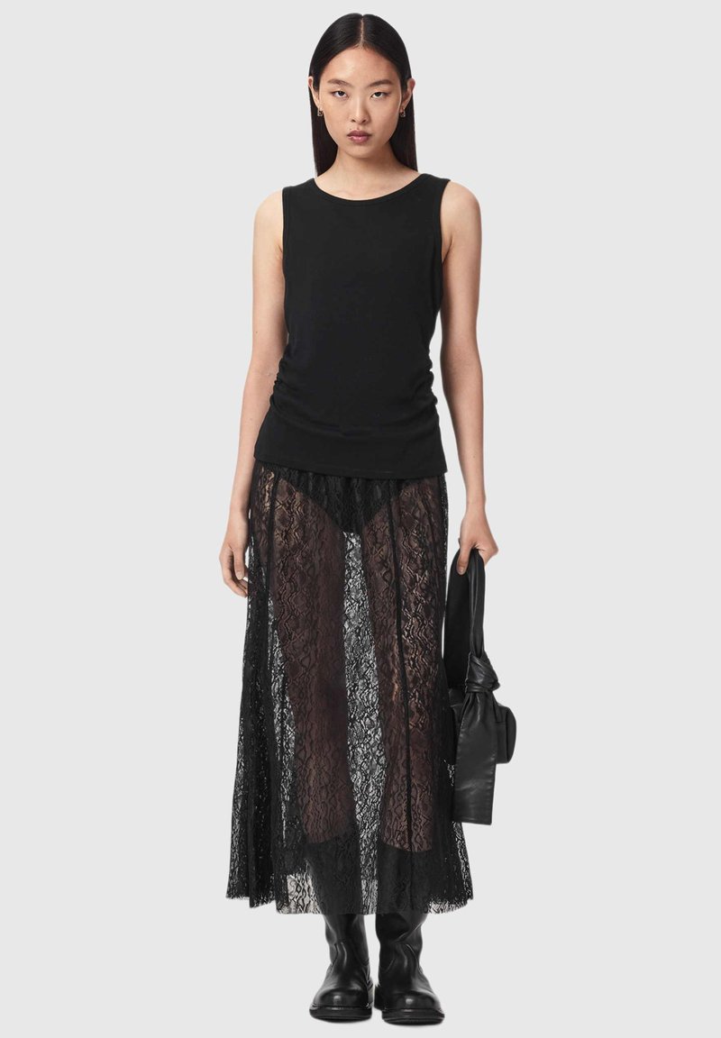 Woman stands facing forward, wearing a black sleeveless top, sheer black lace skirt, black boots, holding a black handbag, neutral background.